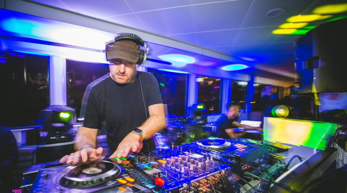 A blurry action shot of a man DJing in a room with lights and CD-J equipment.