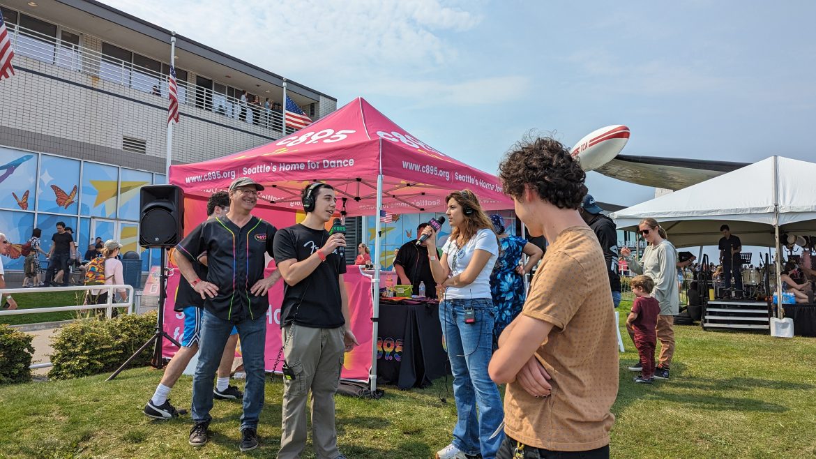 Students on a grass standing in front of a tent with microphones and broadcasting equipment.