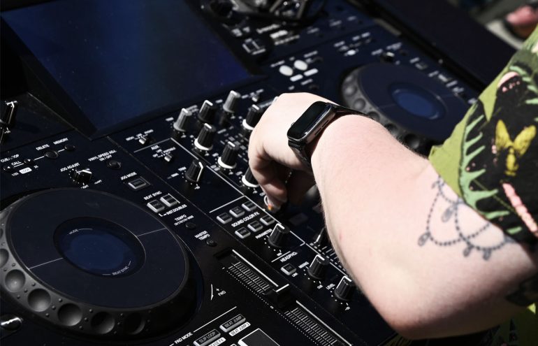 A person adjusting controls on a professional music mixing console, focusing on various knobs and sliders. Their wrist displays a smartwatch, and a tattoo is visible on their arm. The equipment is detailed and technical, suggesting a DJ setup.