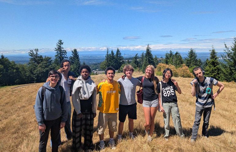 A group of people stands on a grassy hilltop with evergreen trees and a distant city skyline visible in the background under a clear blue sky.